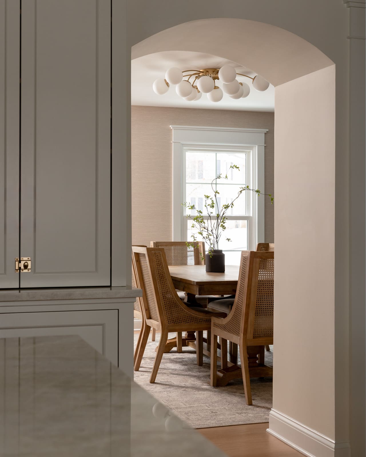 Arched entry from kitchen to dining room with wood table, cane chairs, and bright window.