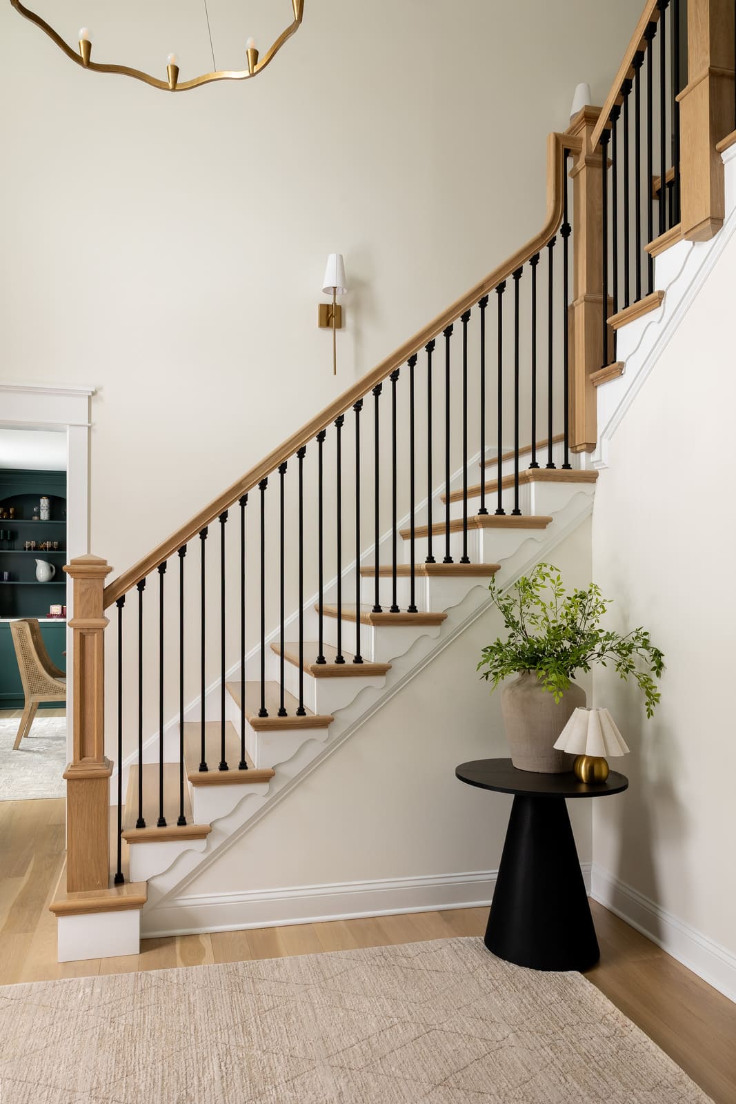 Entryway staircase with wood handrail, black balusters, brass lighting, and bright neutral walls.
