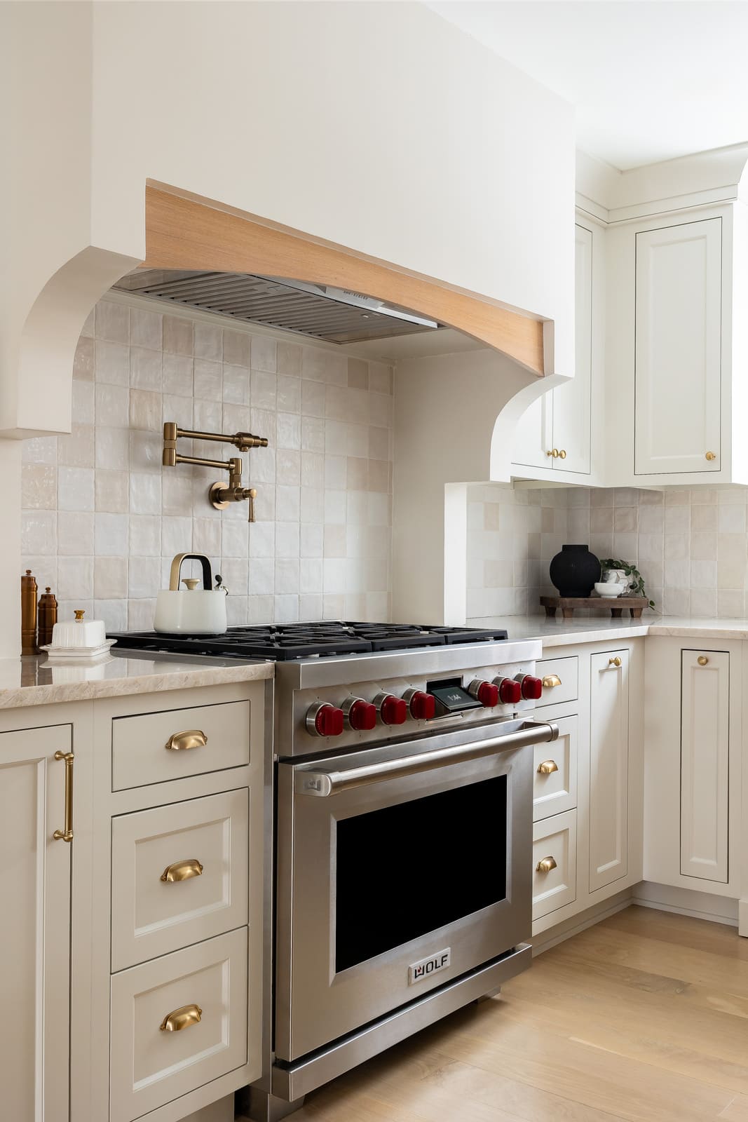 Kitchen range with cream tile backsplash, custom hood, brass pot filler, white cabinets, and wood trim.
