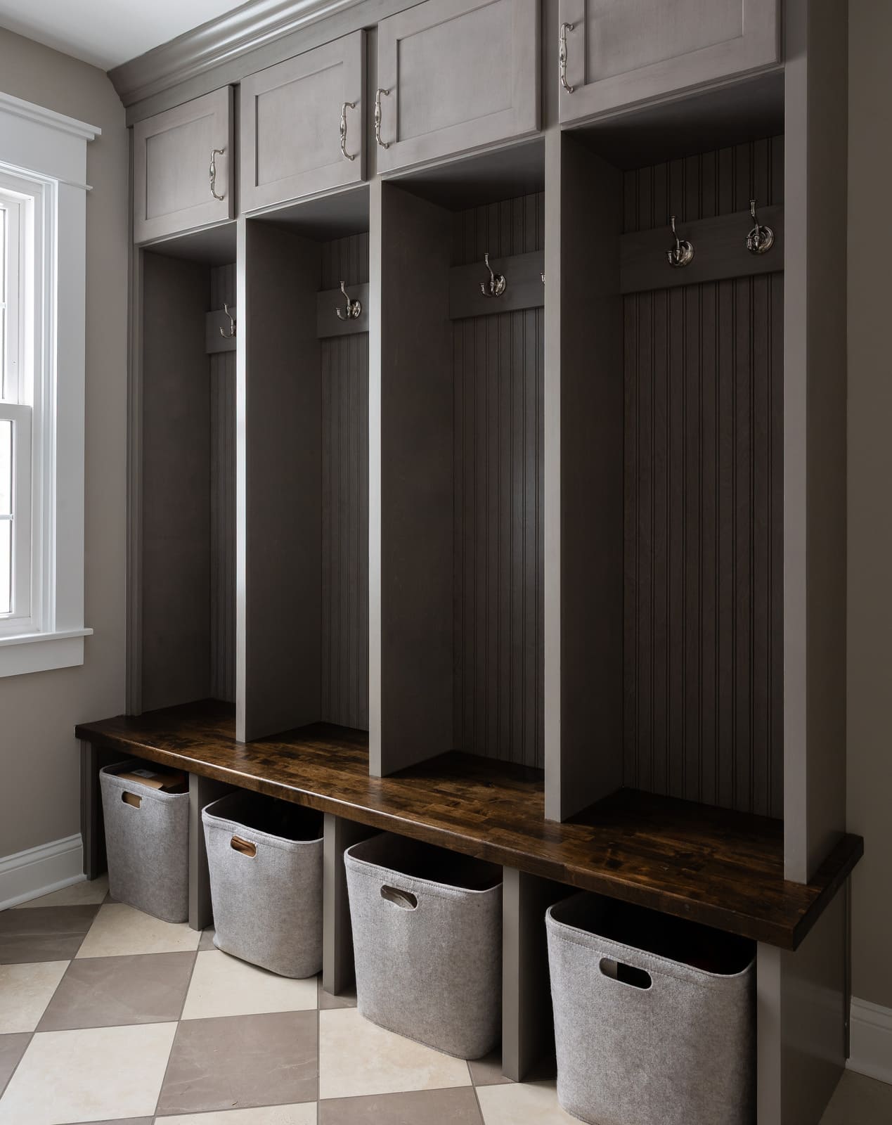 Mudroom remodel with gray built-in cubbies, wood bench, hooks, storage bins, and checkerboard tile floor.