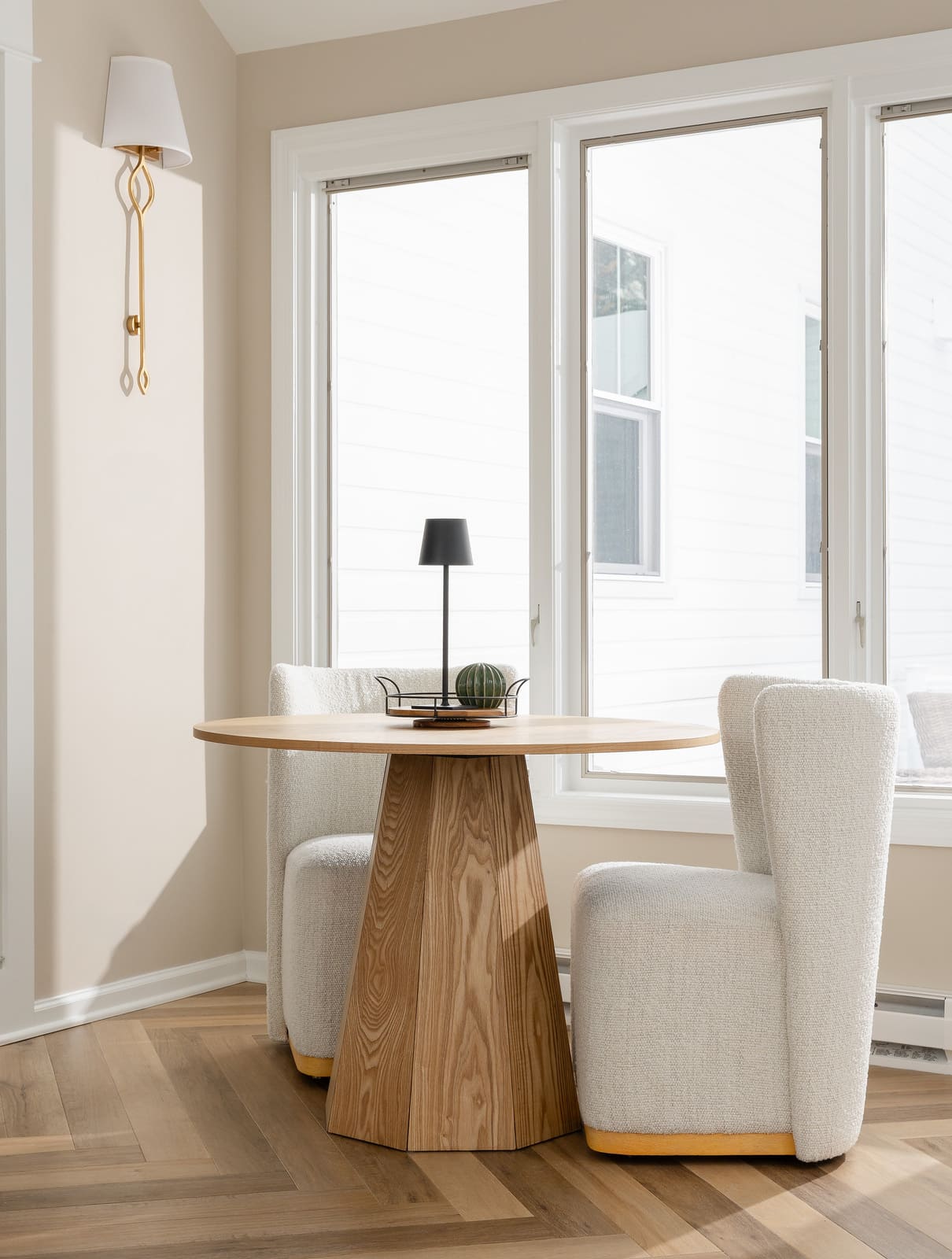 Sunny breakfast area with round wood table, boucle chairs, tall windows, herringbone floor, and brass wall sconce.