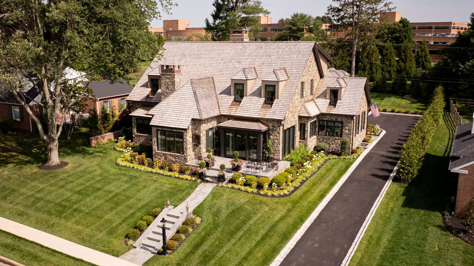 Aerial daytime view of expansive stone Tudor home with manicured lawn, flag, and driveway