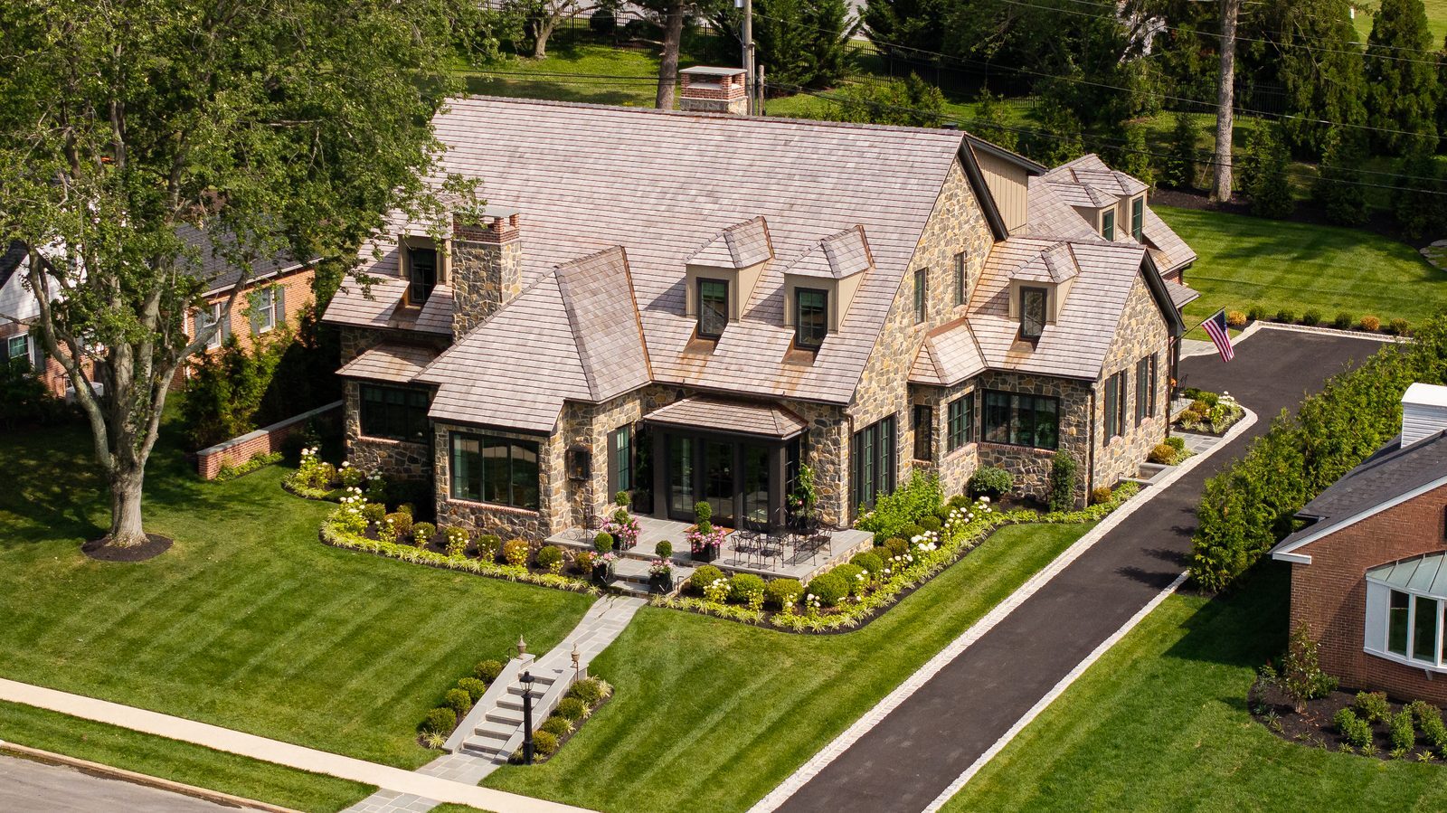Aerial daytime view of stone Tudor home across front lawn with slate roof and trees