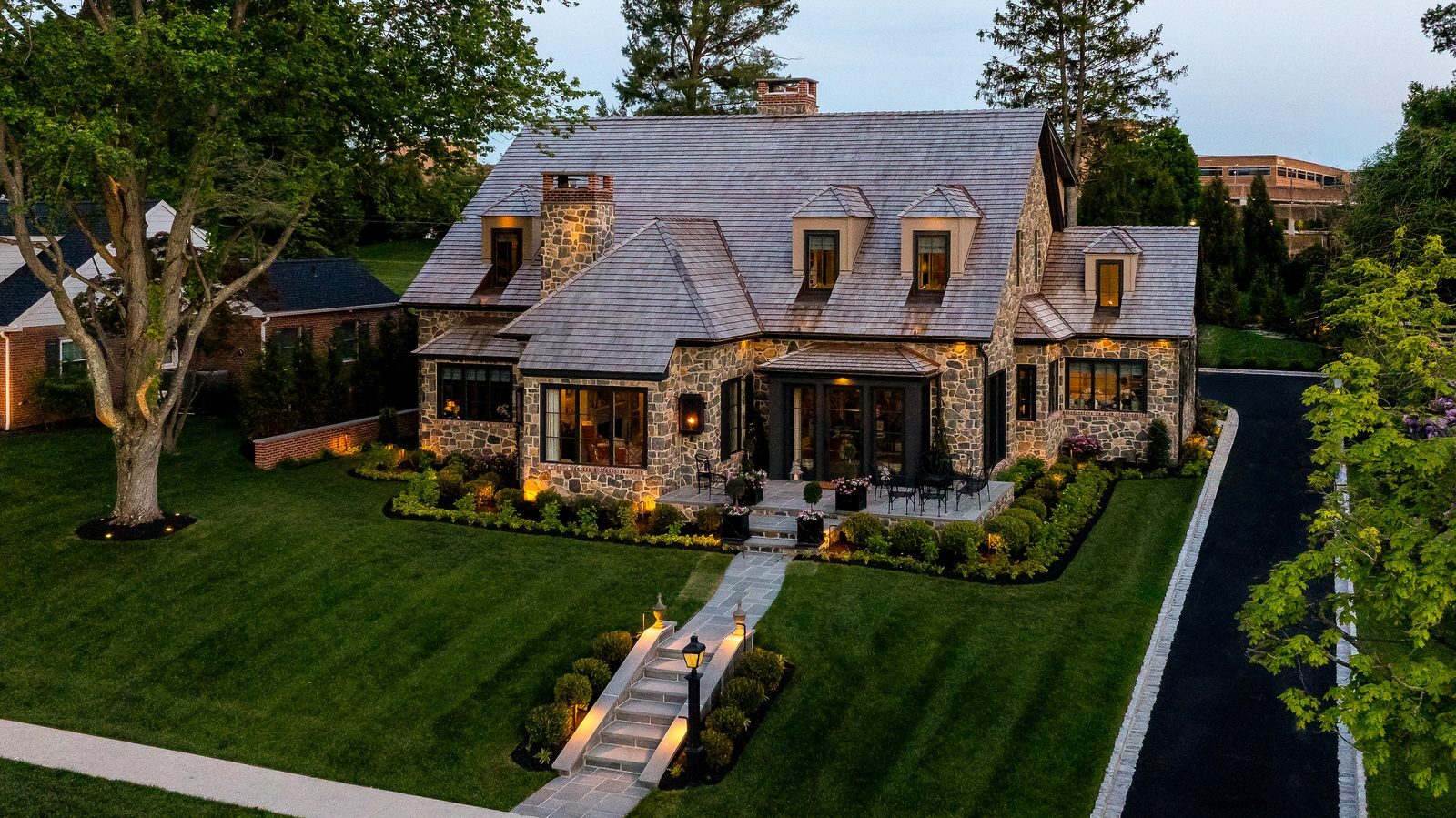 Aerial dusk view of stone home under blue sky with lawn, driveway, and flag