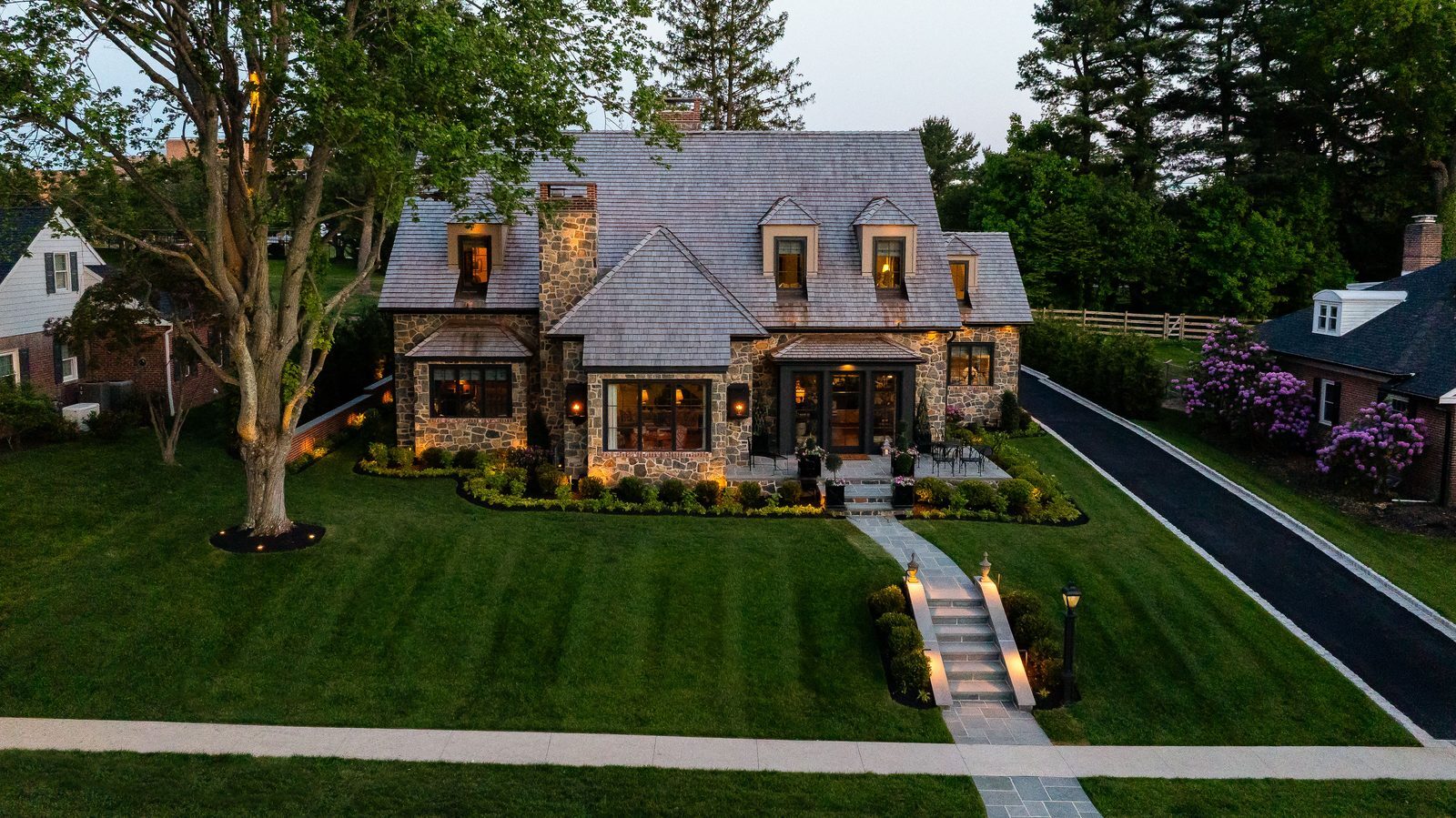 Aerial dusk view of stone home with blooming purple hydrangeas and shade tree