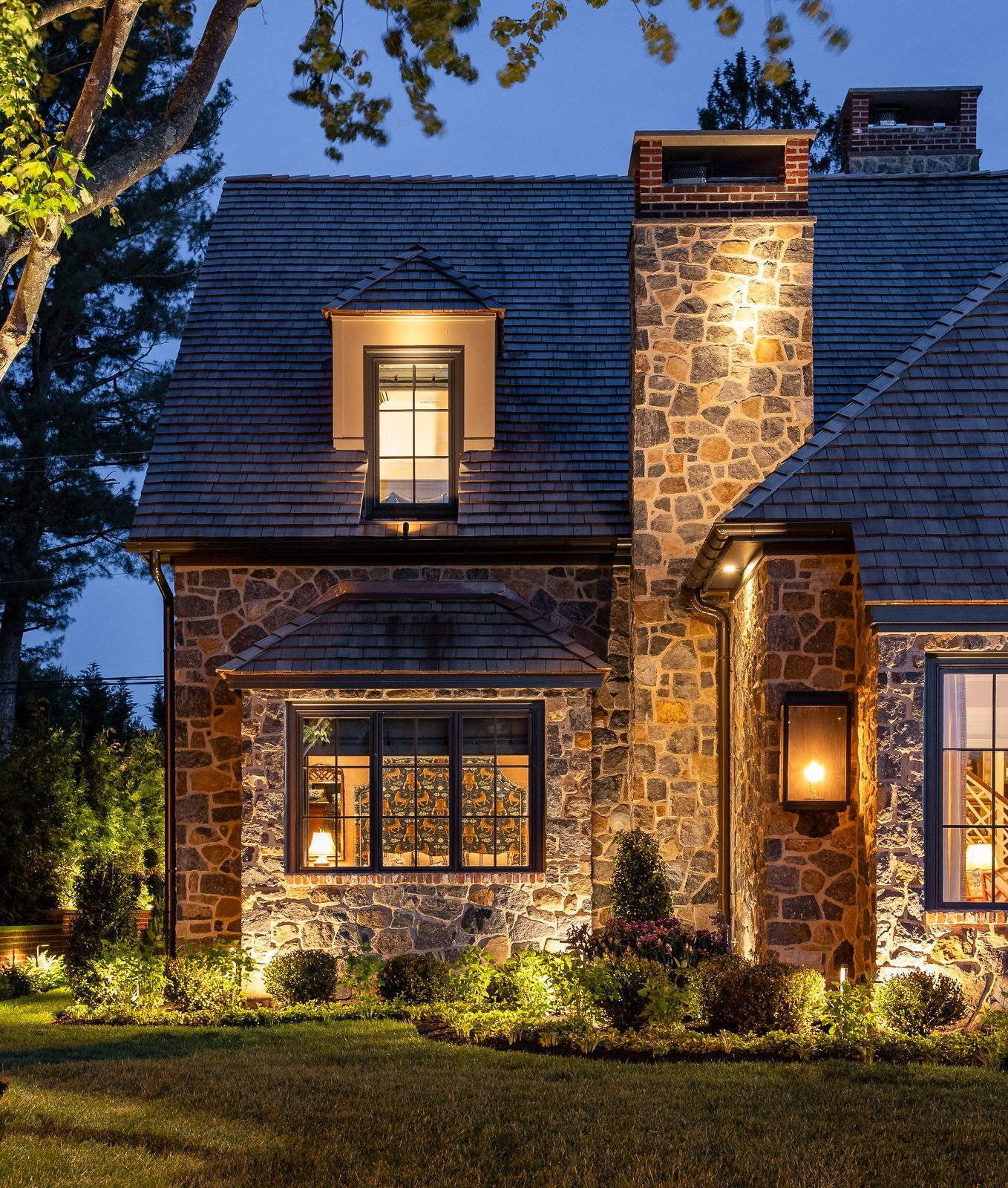 Close-up dusk view of lit dormer window and stone chimney with patterned curtains