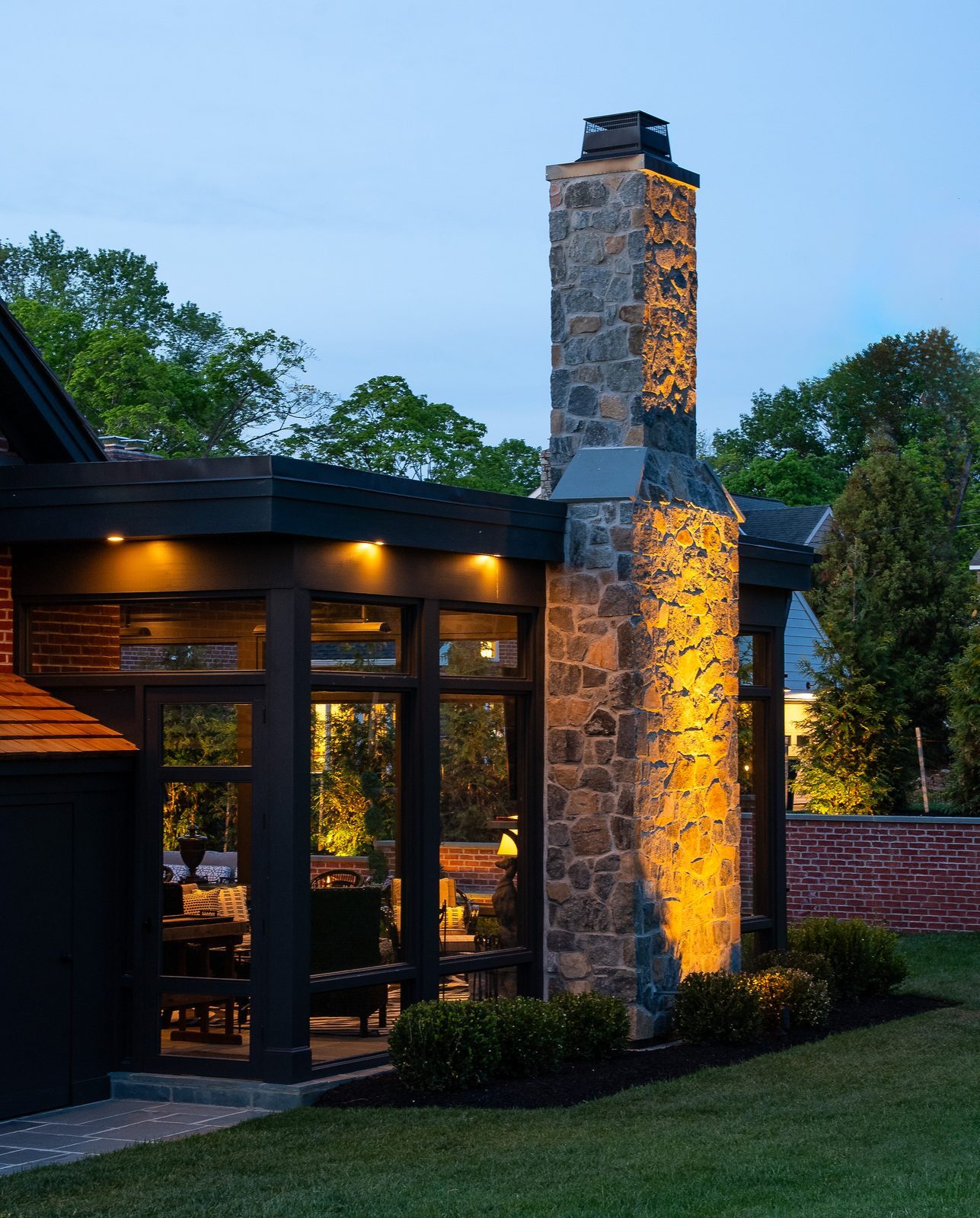 Dusk view of tall stone chimney beside glass sunroom with cozy lit interior