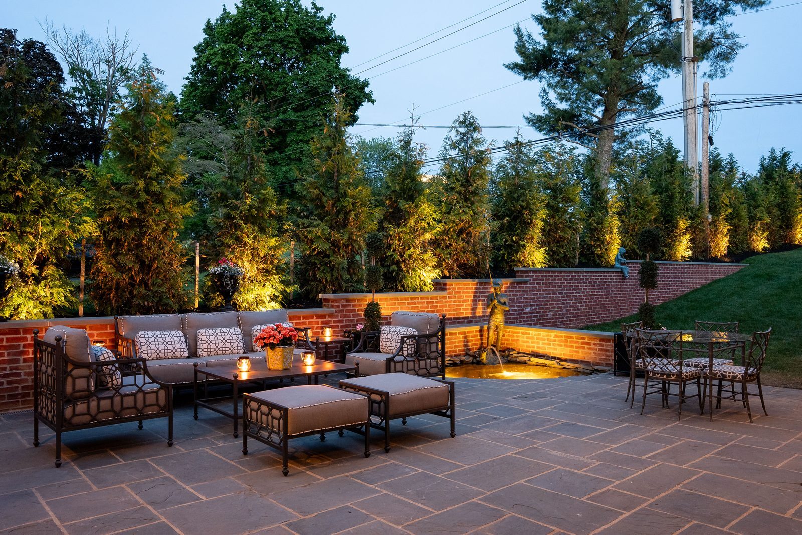 Wide dusk view of backyard patio lounge framed by tall, uplit evergreen trees