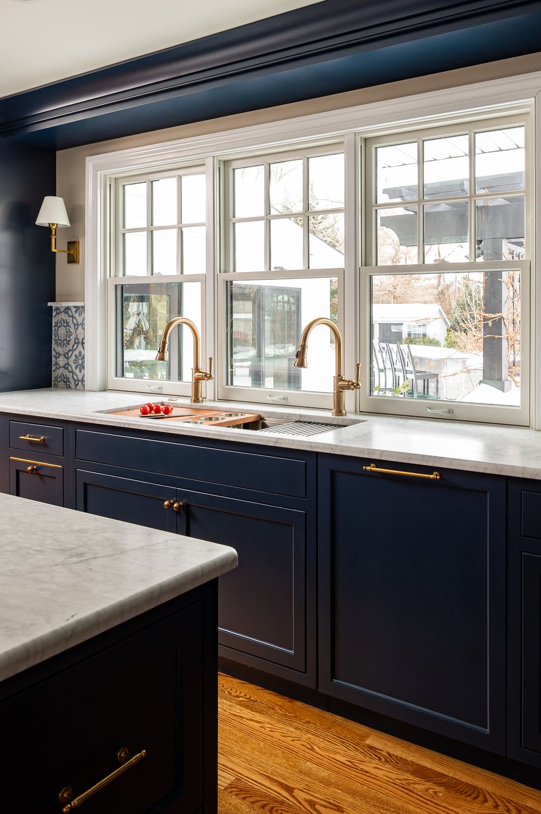 Bright kitchen sink wall with navy cabinets, marble counters, brass faucets, and large windows.