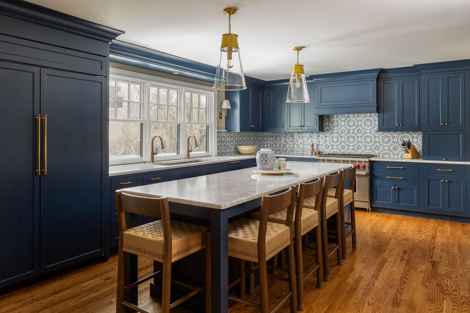 Bright navy kitchen with long marble island, brass pendant lights, patterned tile, and window sink wall. Bright navy kitchen with long marble island, brass pendant lights, patterned tile, and window sink wall.