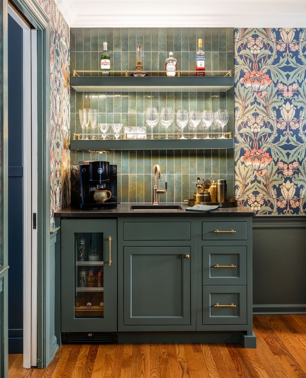 Home bar with green cabinets, dark countertop, brass shelving, floral wallpaper, and vertical green tile.