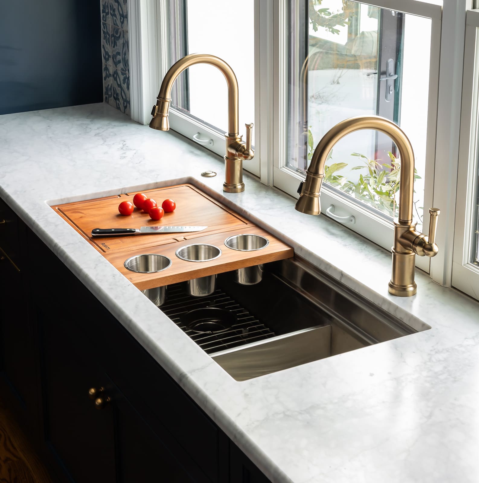 Kitchen sink detail with marble countertop, brass faucets, wood cutting board, and large windows.