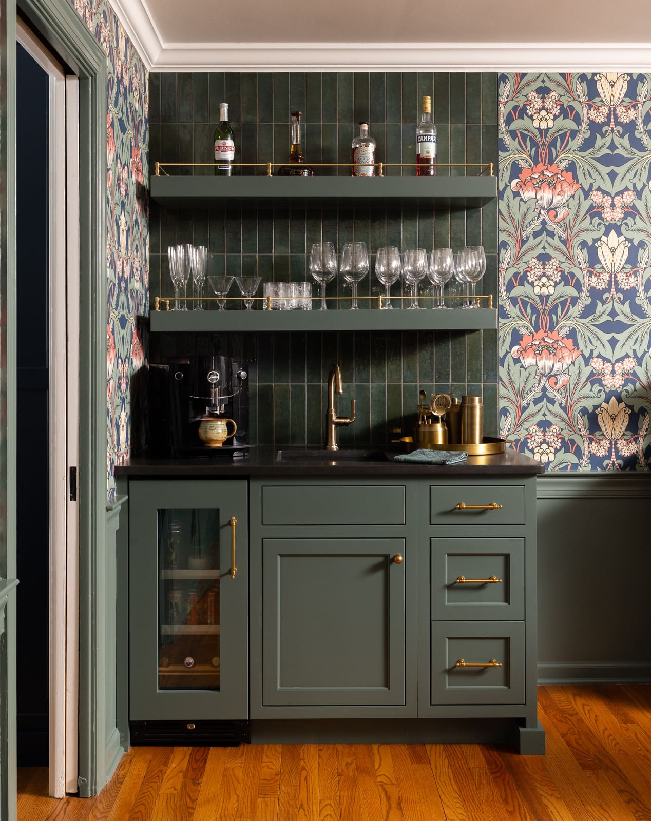 Moody home bar with green cabinetry, brass hardware, glassware shelves, floral wallpaper, and tile backsplash.