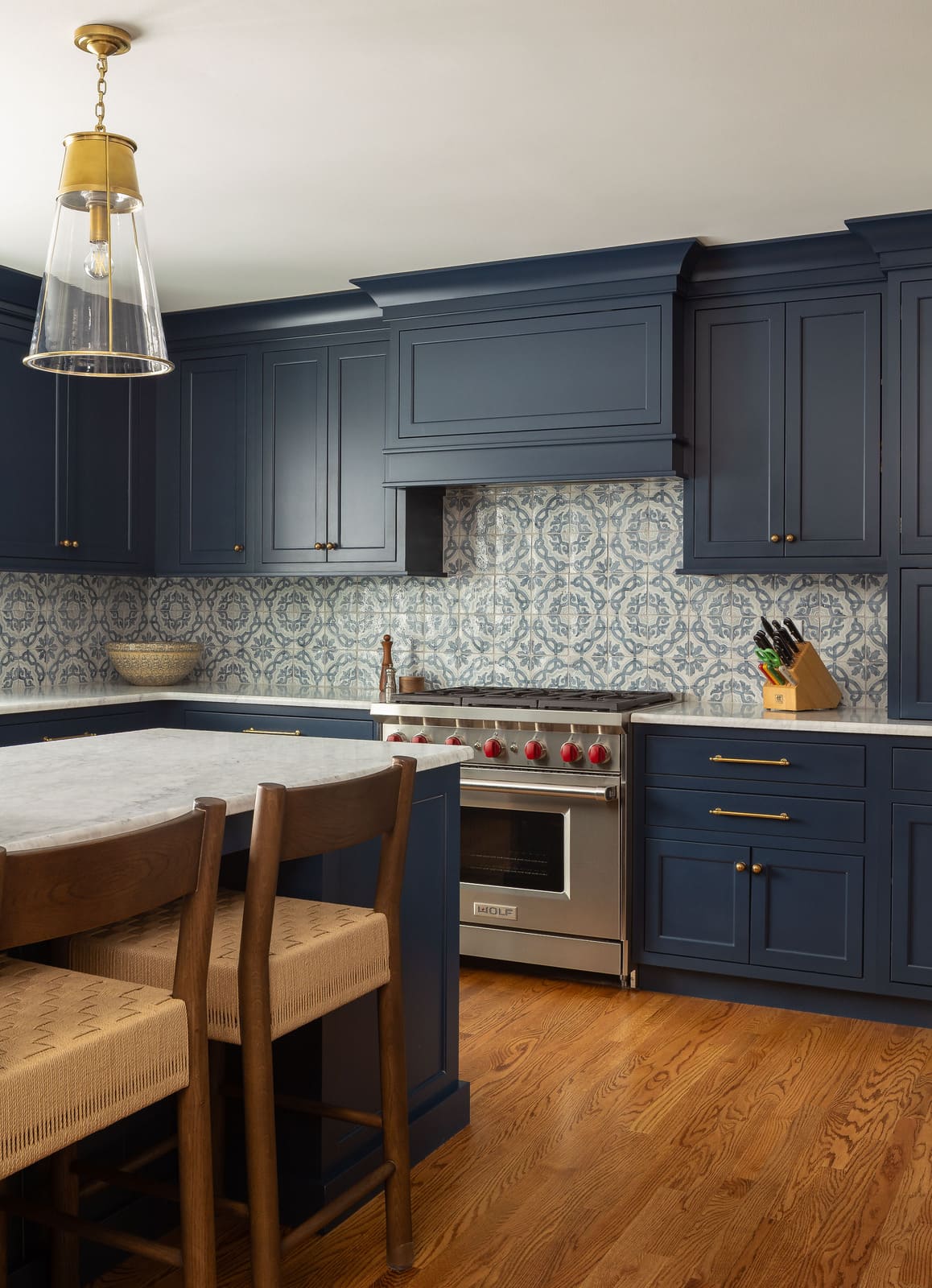 Navy kitchen cabinets with patterned tile backsplash, brass pendant light, marble island, and Wolf range.