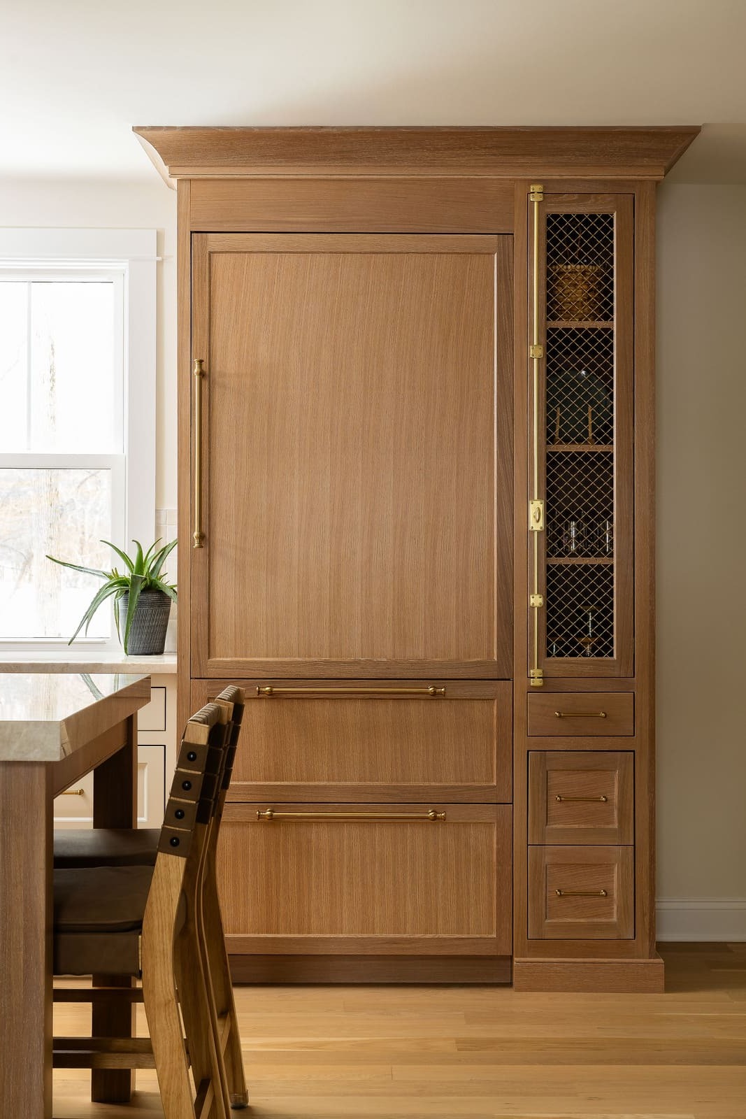 Kitchen storage wall with wood panel refrigerator, brass hardware, glass cabinet, and built-in drawers. Cropped.