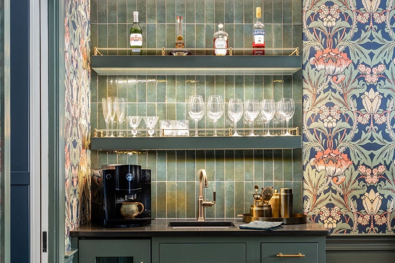 Home bar with green cabinets, dark countertop, brass shelving, floral wallpaper, and vertical green tile